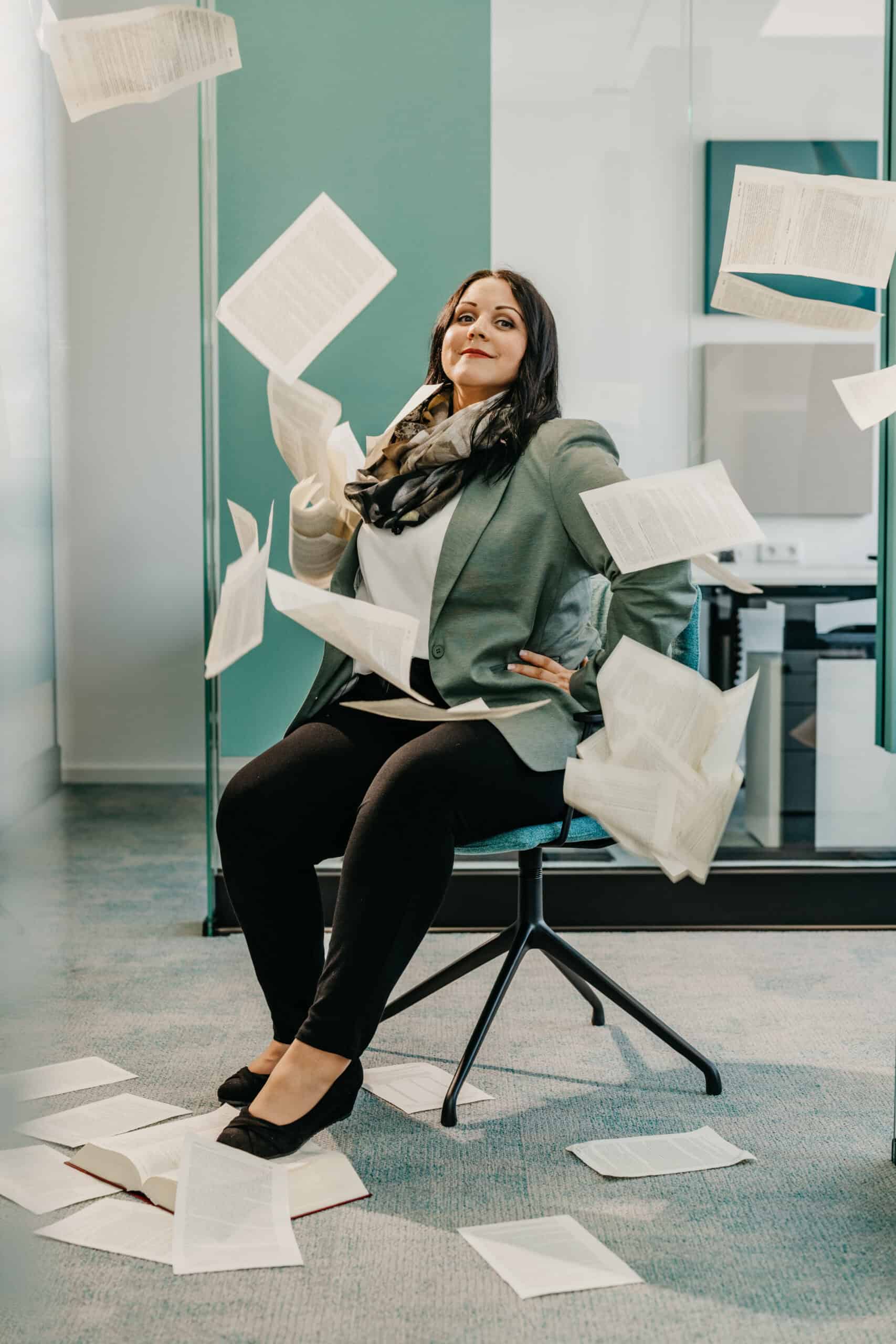 Frau im Büro sitzt auf Stuhl, umgeben von fallenden Papieren und Dokumenten.