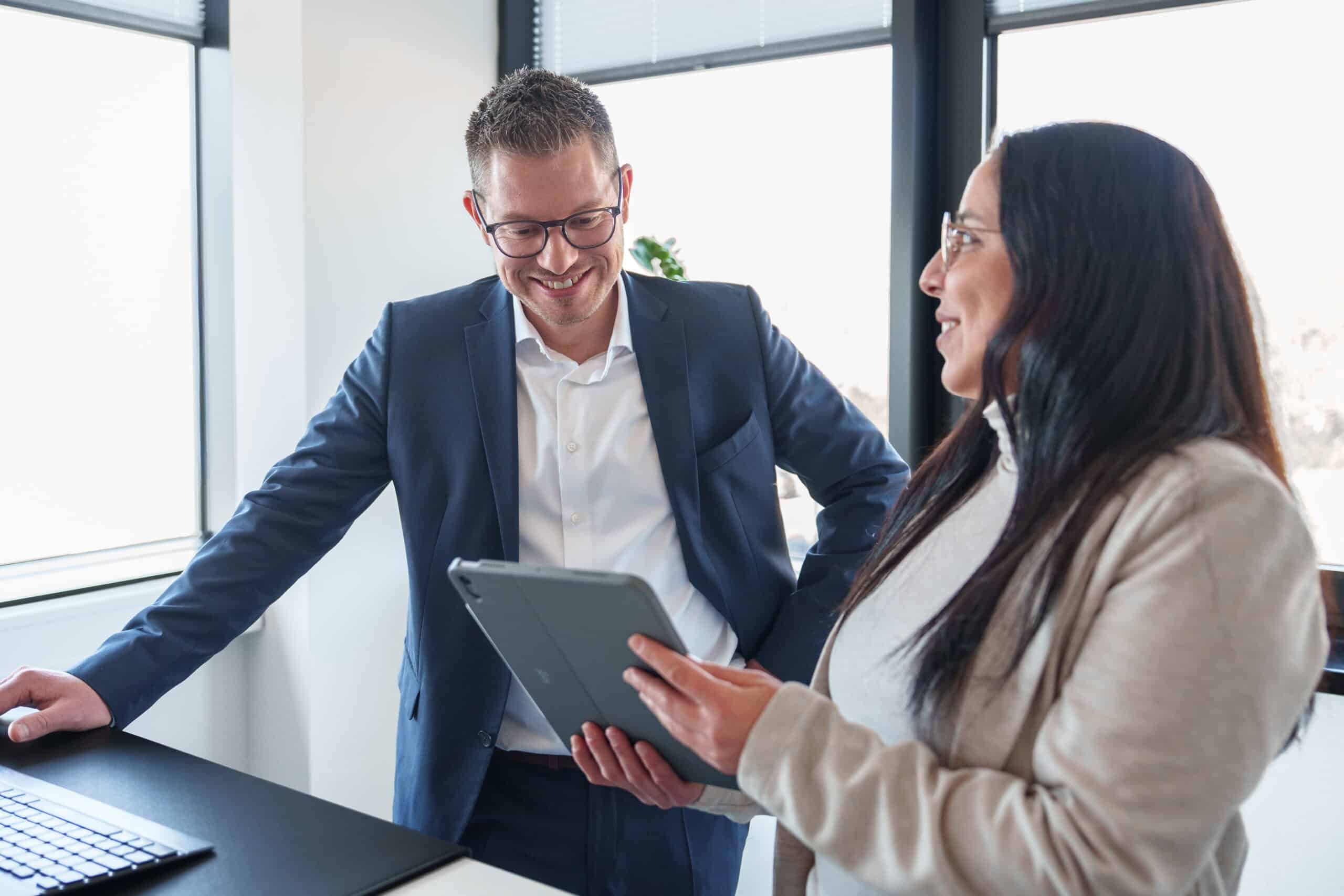 Zwei Geschäftsleute im Gespräch, einer zeigt auf ein Tablet, im modernen Büro mit großen Fenstern.