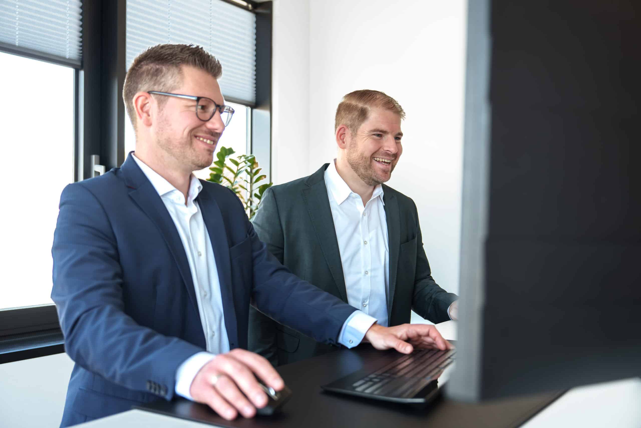 Zwei Männer im Business-Outfit arbeiten gemeinsam am Computer im modernen Büro.